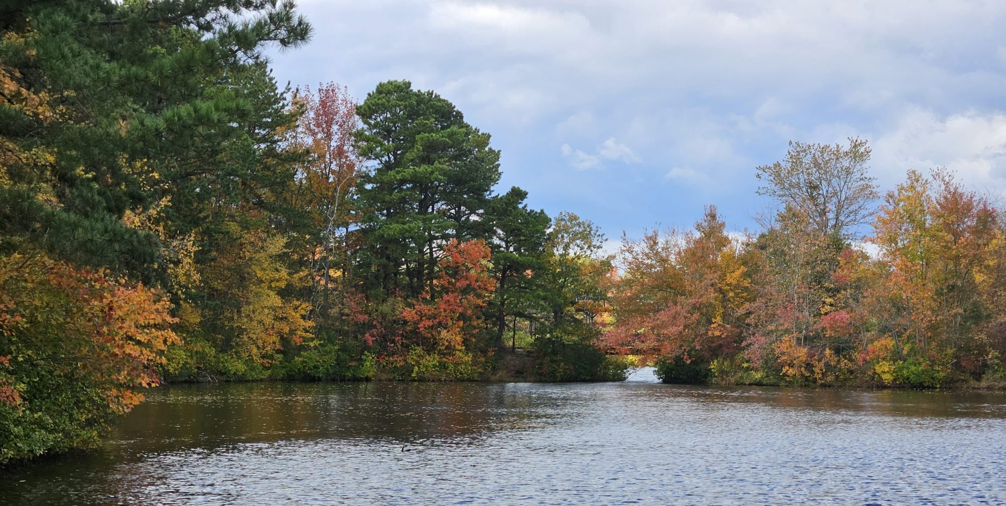 A calm lake surrounded by autumn trees in red, orange, yellow, and green under a cloudy sky.